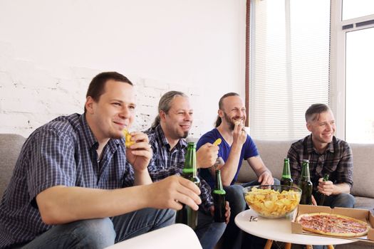 Group of men drinking beer, eating pizza, talking and smiling while resting at home on couch behind TV