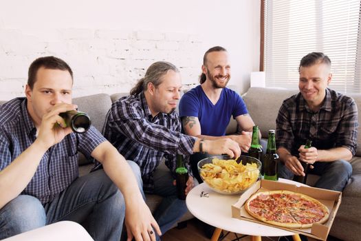 Group of men drinking beer, eating pizza, talking and smiling while resting at home on couch behind TV