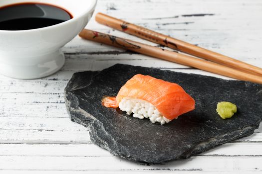 Smoked salmon Nigiri with wasabi paste on slate stone. Chopsticks and bowl with soy sauce in the background on old white wood. Raw fish in traditional Japanese sushi style. Horizontal image.