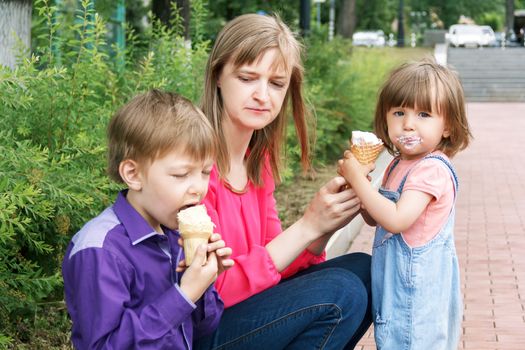 Mother with boy and baby girl sitting in summer park eating icecream