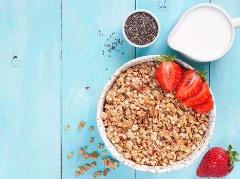 Top view of bowl with muesli, fresh strowberry, chia seeds and milk in milk jug on blue wooden background. Copy space.
