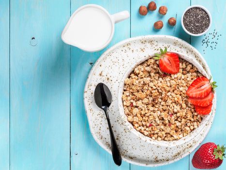 Top view of bowl with muesli, fresh strowberry, nuts, chia seeds and milk in milk jug on blue wooden background. Copy space.