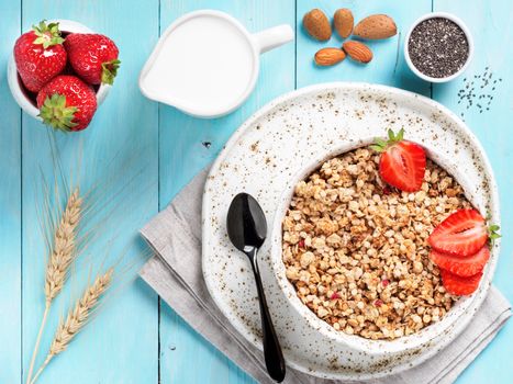 Top view of bowl with muesli, fresh strowberry, nuts, chia seeds and milk in milk jug on blue wooden background. Copy space.