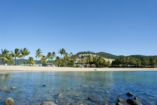 Tranquil view of Airlie Beach, Queensland, Australia across a rocky shoreline and calm ocean to the tropical beach with palm trees and resort buildings