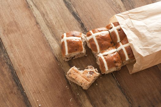 Brown paper packet with fresh hot cross buns from the bakery to celebrate Easter spilling out onto a wooden table with copy space