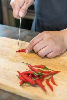 Hand slicing Chilli pepper with Knife on chopping board on wooden background.