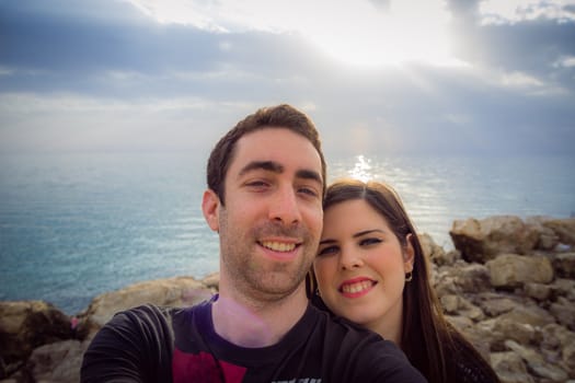 Happy couple taking selfie with smartphone or camera over sunset coast background.