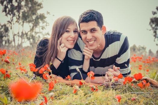 Young couple lying on the grass in a field of red poppies and smiling at the camera.