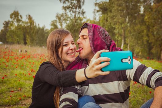 Couple doing silly and funny faces while taking selfie picture with their mobile phone in field of red poppies.