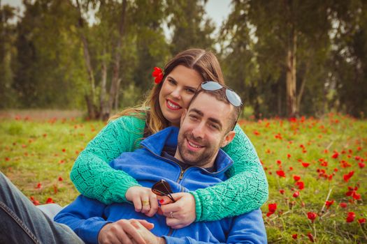 Young couple sitting on the grass in a field of red poppies and smiling at the camera.