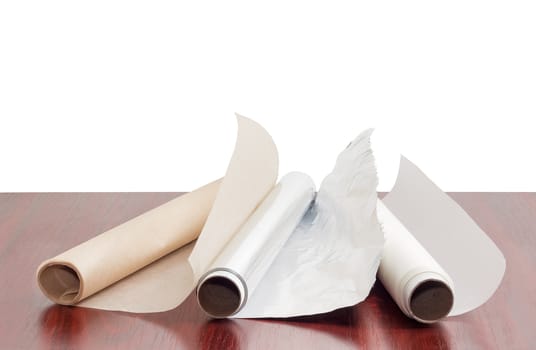 Two rolls of the various parchment paper and one roll of the aluminum foil for household use on a dark red wooden table on a white background
