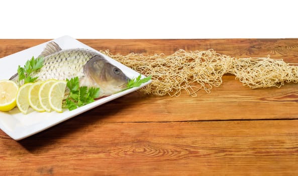 Fragment of the white square dish with carp with peeled scales and lemon slices and parsley on a surface of an old wooden planks with part of the fishing net
