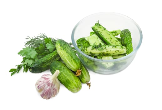 Lightly salted cucumbers in the glass bowl and several freshly picked out cucumbers, parsley, dill and garlic beside on a white background
