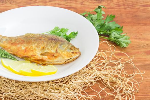 Fragment of the white dish with fried crucian, slices of the lemon, twigs od the parsley and dill and part of the fishing net on a surface of the old wooden planks
