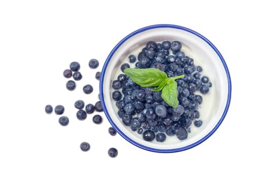 Top view of the blue bowl with dessert made of the fresh blueberries and sweetened condensed milk decorated with basil leaf against of several berries beside on a white background
