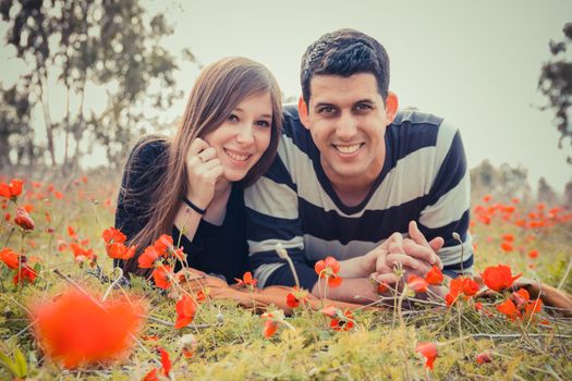 Young couple lying on the grass in a field of red poppies and smiling at the camera.