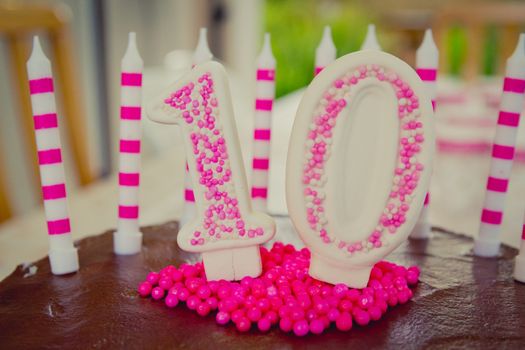 Cake decoration in the figure of the number 10, made of sugar dough and candy in white and pink.
The decoration placed on brown chocolate cake.
There are also 10 candles on the cake.