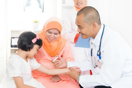 Doctor pediatrician injecting vaccine to Southeast Asian girl. Muslim family.