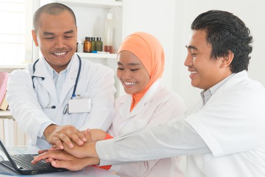 Successful medical doctors stacking hands celebrating inside hospital room. Southeast Asian Muslim medical people.