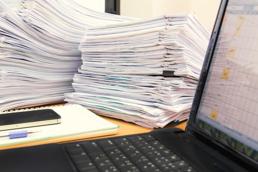 computer notebook and documents on desk stack up high waiting to be managed.