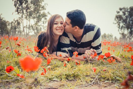 Man kissing woman and she has a toothy smile while they laying on the grass in a field of red poppies.