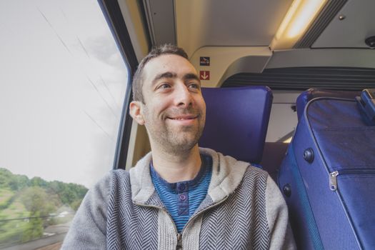 Young man traveling on a train and smiling at someone. A blue suitcase is next to him.