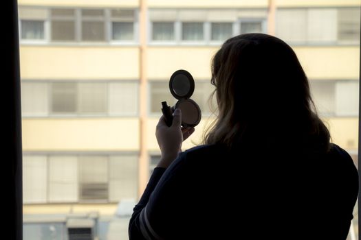 Back view of silhouette of a woman putting on makeup in front of a window.