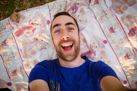 Young man lying on a sheet in the park and taking selfie picture with goofy smile.