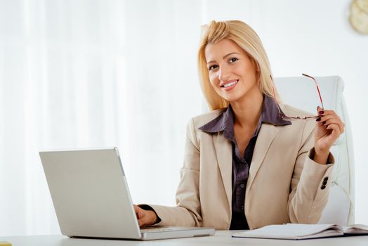 Portrait of a beautiful young businesswoman sitting in the office and smiling. She is holding glasses and looking at camera. 