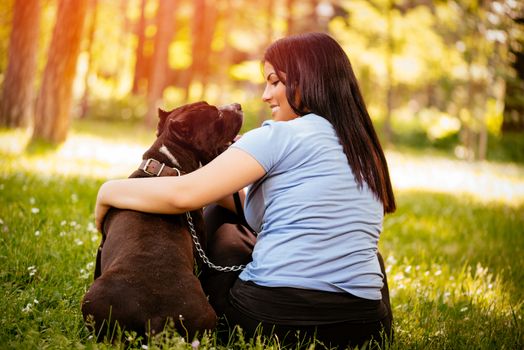 Rear view of a beautiful young woman hugging her cute stafford terrier. They sitting on grass in the park and enjoying. 