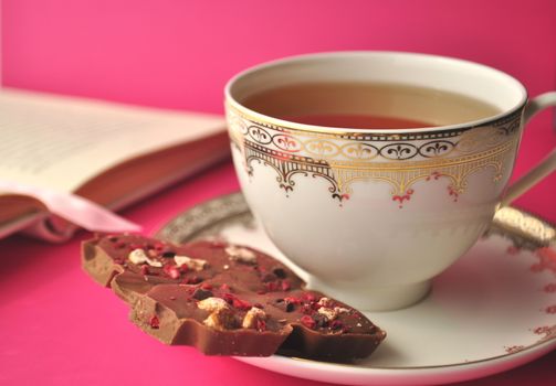 Cup of tea with chocolate and a book on bright pink background with copy space