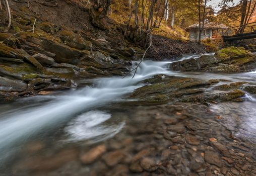 Autumn view of the flow water of a small river with a bridge and a house in the mountains