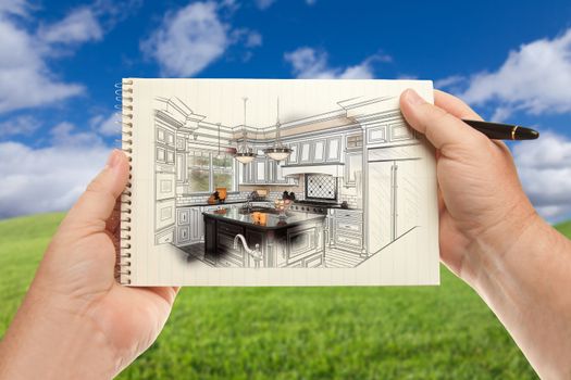 Male Hands Holding Pen and Pad of Paper Up with Custom Kitchen Illustration Outside Near Grass Field and Blue Sky.