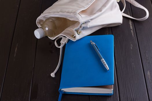 White leather backpack, blue notebook and pen on a black wooden background