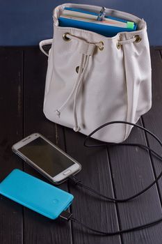 white smartphone is charged from a powerbank near a backpack on a black wooden background