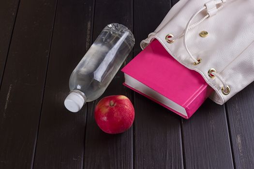 White leather backpack, pink book, bottle of water on a black background