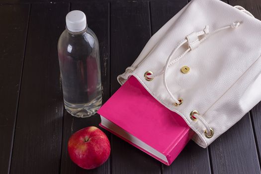 White leather backpack, pink book, bottle of water on a black background