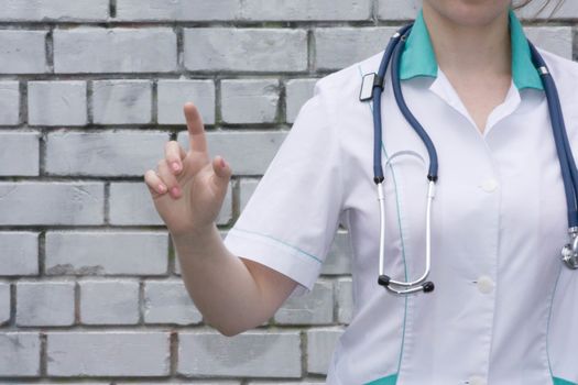 The doctor's concept. Girl in medical uniform with a stethoscope near a brick wall. Holds a finger finger forward. Presses. Photo for your design