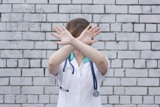 The doctor's concept. Girl in medical uniform with a stethoscope near a brick wall. Holds his hands at the face, overlapping the cross. Photo for your design