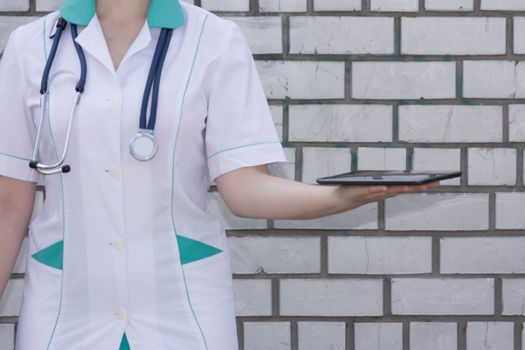 The doctor's concept. Girl in medical uniform with a stethoscope near a brick wall. Holds a tablet in his hand. Photo for your design