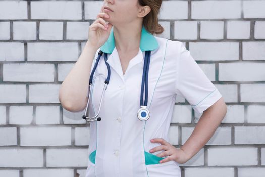 The doctor's concept. Girl in medical uniform with a stethoscope near a brick wall. Meditation. With a hand under his chin. Photo for your design