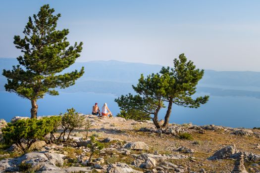 A mature couple sitting on a large rock under some trees, overlooking the seashore at sunset and looks at the horizon and the sea