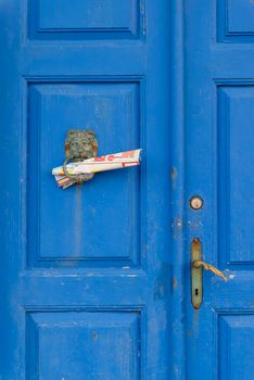 A vintage blue wooden double door with bronze knocker and newspapers attached to knocker
