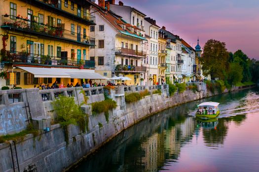 Panorama of old town Ljubljana, Slovenia, with numerous bars and restaurants at waterfront of Ljubljanica river in sunset, panorama boat on river, clouds reflection in water