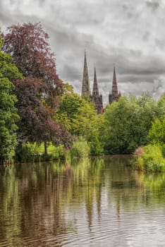 A view of the three spires of Lichfield cathedral taken from the local park showing reflections in the pool