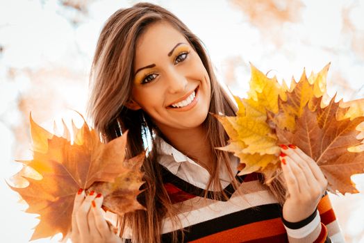 Portrait of a cute young woman enjoying in sunny forest in autumn colors. She is holding golden yellow leaves. Looking at camera.