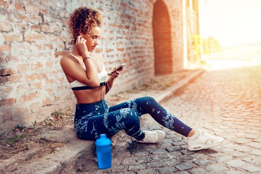 Young sporty woman with headphones taking a break after hard training and using mobile phone.
