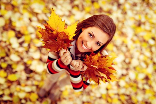 Beautiful young woman enjoying in sunny forest in autumn colors. She is holding golden yellow leaves. Looking at camera. Top view.