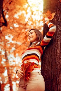 Beautiful young woman enjoying in sunny forest in autumn colors. She is holding golden leaves and standing next to tree. 