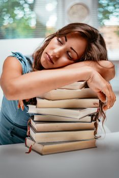 Tired teenage girl sitting in library and sleeping with hands on stack of books. 